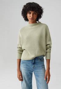 Hellgrüner gerippter Pullover mit hohem Kragen, vollem Ärmeln und lässiger Passform, kombiniert mit hellblauen geraden Jeans.