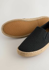 Alpargatas negras tipo slip-on con empeines de tela texturizada, medias suelas trenzadas de yute y suelas exteriores de goma con patrón de guijarros, exhibidas sobre una superficie neutra.