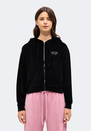 Femme portant un sweat à capuche zippé noir avec le texte "Moschino Active" et un pantalon à cordon rose clair, debout devant un fond uni.