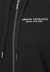 Černá mikina s zipem vyrobená z hladkého materiálu. Obsahuje stříbrný zip, nastavitelné šňůrky a bílé logo "Armani Exchange" na hrudi.