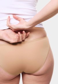 Slip beige senza cuciture con una texture liscia, caratterizzati da un design a vita bassa e cuciture minime, visti di spalle con le mani sui fianchi.