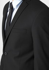 Czarna marynarka o pojedynczym guzie, z klapami w kształcie notcha oraz lewą kieszenią na piersi. Charakteryzuje się gładką teksturą i dopasowanym krojem. Pod spodem biała koszula.