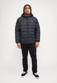 Black puffer jacket with a hood, quilted design, and front zipper. Paired with black pants and black sneakers with white laces.