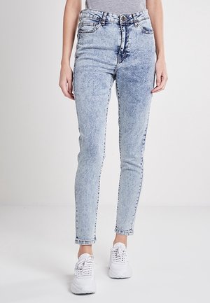 Jean skinny taille haute bleu clair porté avec des baskets blanches et un haut gris, présenté de la taille aux pieds sur un fond neutre.