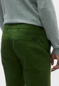 Πράσινα βαμβακερά chinos με ευθεία γραμμή, διαθέτουν πίσω τσέπες και ετικέτα λογότυπου. Φοριούνται με γκρι πουλόβερ, παρουσιάζοντας καθαρές γραμμές και απαλή υφή.