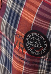 Camicia di tessuto a quadri con linee rosse, blu e bianche; presenta una toppa rotonda nera con un design di montagna e la scritta "Vancouver".