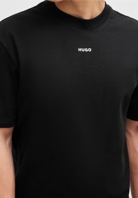 Czarna koszulka z bawełny z krótkimi rękawami, z okrągłym dekoltem i białym logo "HUGO" nadrukowanym na piersi.