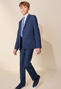 Niño con traje azul a cuadros, camisa azul clara y corbata azul, de pie con una pierna ligeramente adelantada sobre un fondo liso.