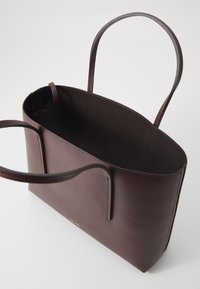 Sac fourre-tout en cuir bordeaux avec deux longues poignées, un intérieur spacieux et un design minimaliste agrémenté d'un logo doré à l'avant.