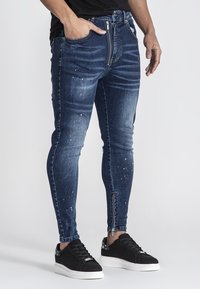 Vaqueros ajustados de color azul con áreas desgastadas y manchas de pintura blanca dispersas, que cuentan con una cremallera y acentos cosidos a lo largo del costado.