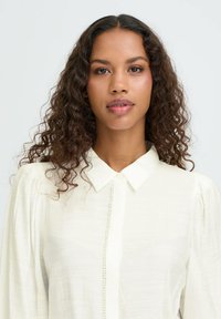 Blusa blanca con botones en la parte delantera, cuello con solapa y mangas abullonadas; hecha de una tela ligera y texturizada.
