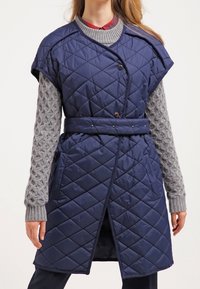 Marinblå kviltad ärmlös kappa med bälte i midjan, med ett diamantmönster och två framfickor. Grått strukturerat tröja under.