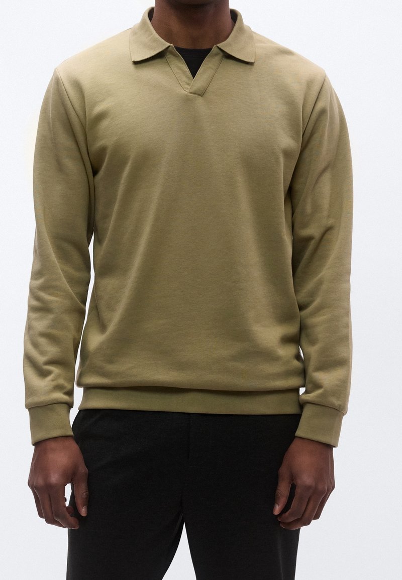 Homme portant un polo manches longues vert olive avec col et pantalon noir sur un fond blanc uni.