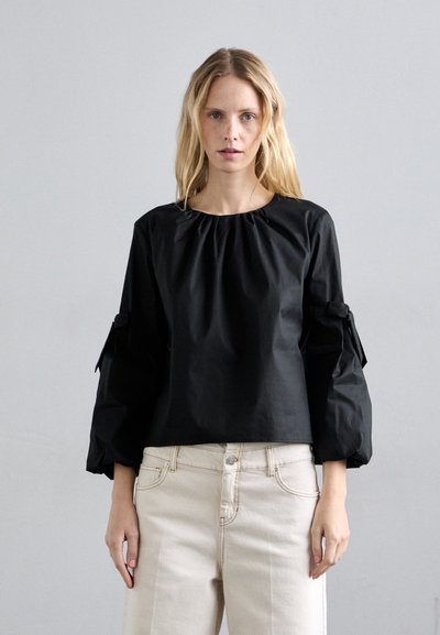 Čierny skrátený blúz s riaseným golierom a objemnými rukávmi, s detailmi viazania po stranách. Skombinované so svetlými džínsami.
