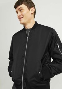 Jachetă bomber neagră dintr-un material neted, cu fermoar frontal, manșete ribbate și buzunare laterale, având un design curat și minimalist.