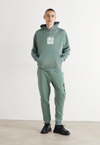 Sudadera verde con el logo "HUGO" y pantalones de chándal a juego, ambos de una tela suave. Unos zapatos negros completan el atuendo casual.