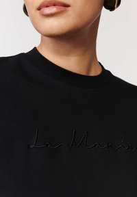 Czarna bluza z okrągłym dekoltem, ozdobiona fakturowanym haftem "La Manina" na klatce piersiowej. Gładki materiał o miękkim wykończeniu.