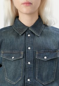 Tumši zili denima krekls ar norādīto apkakli, pogu priekšpusi ar metāla pogām un divām krūšu kabatām ar vākiem.