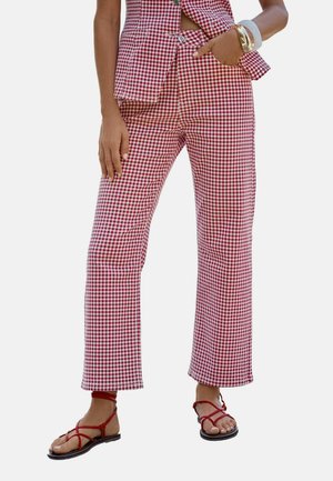 Femme portant un pantalon à carreaux rouges et blancs et un haut sans manches assorti avec des boutons, accessoirisée de bracelets et de sandales rouges à lanières.