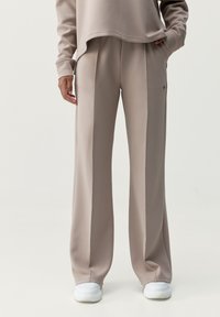 Svetlé béžové kalhoty širokého střihu vyrobené z hladké tkaniny, s předními záhyby a bočními kapsami, doplněné o bílé tenisky.