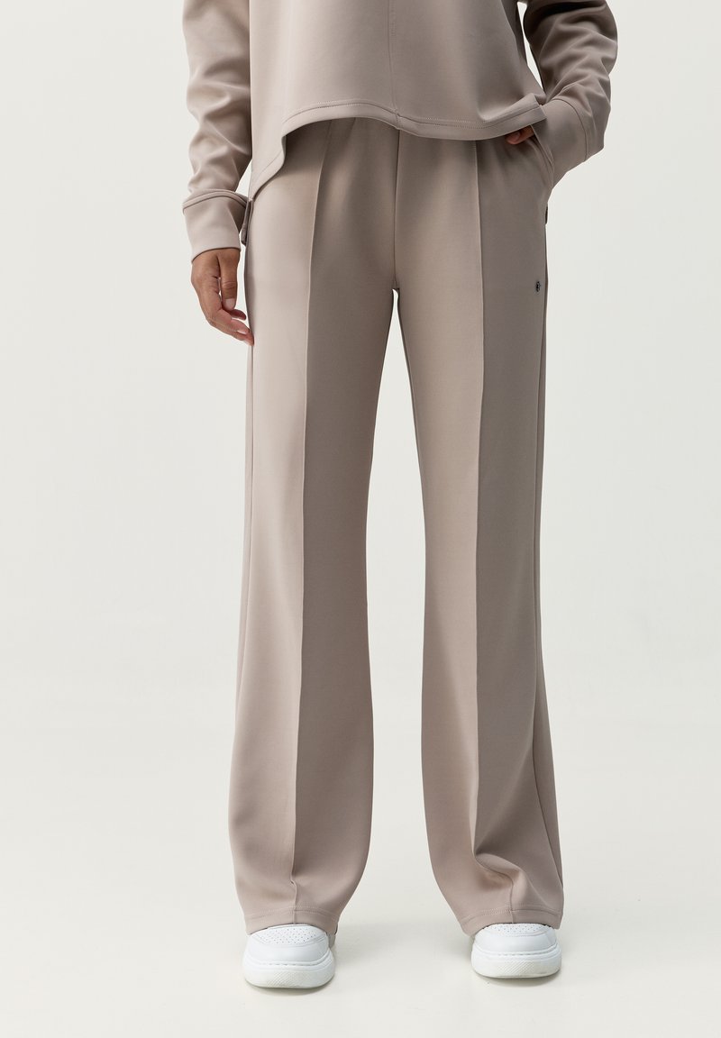 Svetlé béžové kalhoty širokého střihu vyrobené z hladké tkaniny, s předními záhyby a bočními kapsami, doplněné o bílé tenisky.