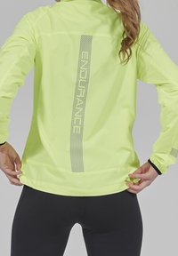 Světlé žluté sportovní sako vyrobené z lehkého materiálu, s reflexními akcenty a přiléhavým střihem pro zvýšenou viditelnost během aktivit.