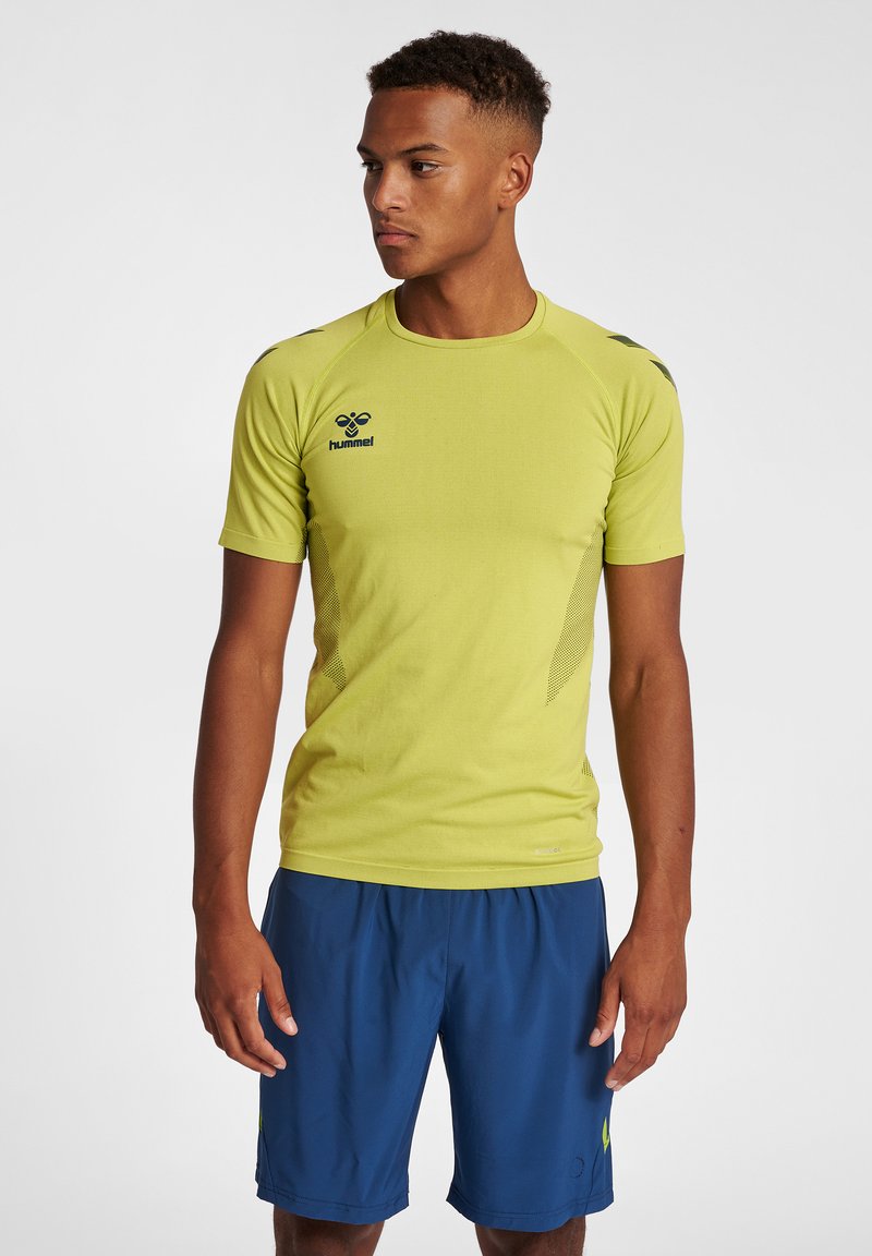 Jonge man met een geel hummel sport-T-shirt en blauwe atletiekshorts staat met ontspannen armen en kijkt naar links tegen een effen achtergrond.