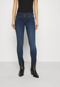 Mujer vistiendo pantalones ajustados de tiro alto azul oscuro con cierre de doble botón y botas de tacón negro hasta el tobillo, contra un fondo liso.