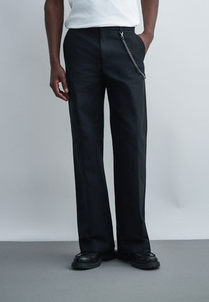 Homme portant un pantalon noir droit avec un détail de chaîne sur le côté, des chaussures noires épaisses et une chemise blanche rentrée, debout devant un fond uni.