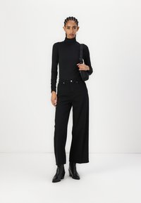 Crna turtleneck majica u kombinaciji s crnim širokim hlačama, izrađenim od glatkog materijala. Model nosi crne čizme na visoku petu do gležnja i nosi torbu.