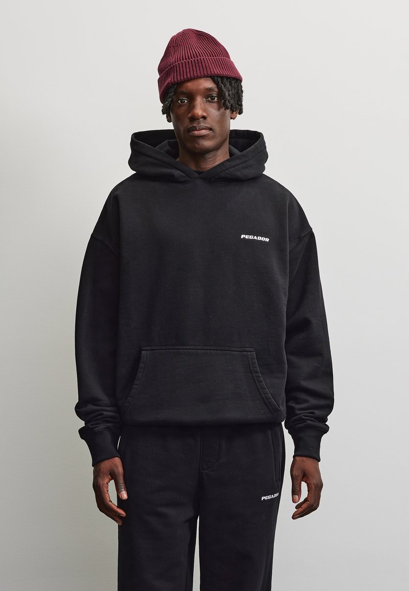 Hombre con sudadera con capucha y pantalones negros de Pegador y un gorro de punto burdeos, de pie frente a un fondo liso gris claro.