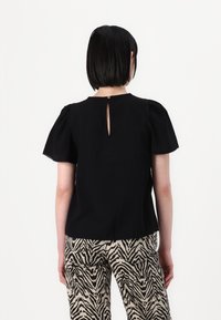 Juoda blusa su puffed rankovėmis, atvira nugara ir banguotais kraštais. Derinama su smėlio ir juodais dryžuotais kelnėmis.