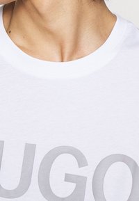 Hvid bomulds t-shirt med rund halsudskæring og et stort gråt "HUGO" logo. Stoffet ser blødt ud med en glat tekstur.