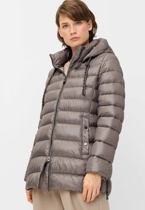 Woman wearing a gray quilted puffer jacket with a high collar and hood, standing against a plain light background.