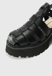 Sandalo in pelle nera con design a strisce, punta aperta, suola in gomma spessa e dettagli intrecciati. Presenta una piattaforma robusta e strutturata.