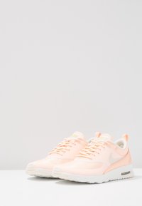 Ett par ljusrosa Nike löparskor med vita sulor och snörningsdesign på en vit yta mot en enkel ljus bakgrund.