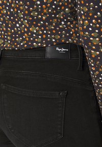 Jeans de algodón negros con un bolsillo trasero y una etiqueta de marca distintiva. La parte superior tiene un patrón floral en naranja, blanco y verde sobre un fondo oscuro.