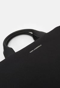 Sort mulepose lavet af glat stof med læderdetaljer. Har afrundede håndtag og en metallisk logodetalje på forsiden.