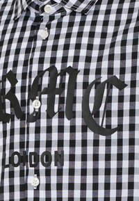 Koszula w czarno-białą kratę z kołnierzykiem na guziki, białymi guzikami i wyraźnym czarnym nadrukiem "KMC LONDON."