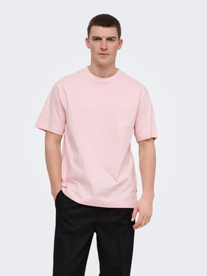 Homme debout, les mains dans les poches, portant un t-shirt rose clair à manches courtes et un pantalon noir sur un fond uni.