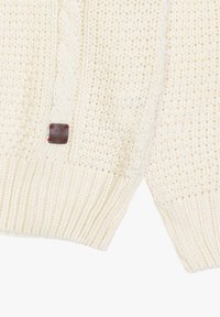 Chunky crèmekleurig gebreid sweater met kabel- en ribbeltexturen, voorzien van een klein bruin leren label aan de zoom. Zacht en warm materiaal.