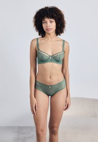 Conjunto de sutiã e cueca em rendas verdes com tecido transparente, bordas em formato de concha e um enfeite central no sutiã e na cinturinha.