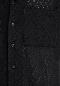 Chemise noire en tricot avec un motif texturé en forme de losange. Comprend une poche avant et des boutons noirs.