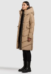 Béžový prošívaný kabát s kapucí a zapínáním na zip vpředu, s diagonálním prošíváním a bočními zipy pro větrání. Modelka má na sobě černé legíny a boty.