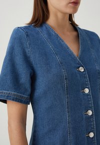 Τζιν φόρεμα σε μεσαίο μπλε απόχρωση, με V-λαιμόκοψη, κοντά μανίκια και μεταλλικά κουμπιά κατά μήκος του μπροστινού μέρους. Υφασμάτινη υφή με ορατές ραφές.