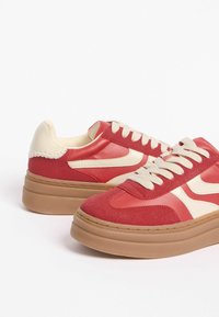 Raudoni sportbačiai su švelniu ir tekstūruotu paviršiumi, turintys kremiškus akcentus ir storą smėlio spalvos padą, užsegami raišteliais ir siuvinėti detaliai.