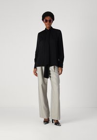 Schwarze Bluse mit langen Ärmeln und Bindedetail, kombiniert mit hellgrauen, gestreiften Hose und schwarzen High Heels. Braune Sonnenbrille.