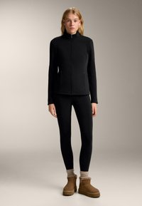 Fekete, cipzáros felső magas nyakrésszel, fekete leggingsszel és barna bokacsizmával párosítva. A ruha sima textúrájú és testhezálló kialakítású.