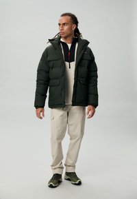 Groene pufferjack met een hoge kraag en meerdere zakken aan de voorkant, gelaagd over een zwart en beige fleece, gecombineerd met beige cargobroeken en groene schoenen.