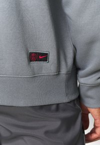 Sweat-shirt gris avec un petit patch logo Paris Saint-Germain et Nike rose près de la taille, porté sur un pantalon gris foncé.